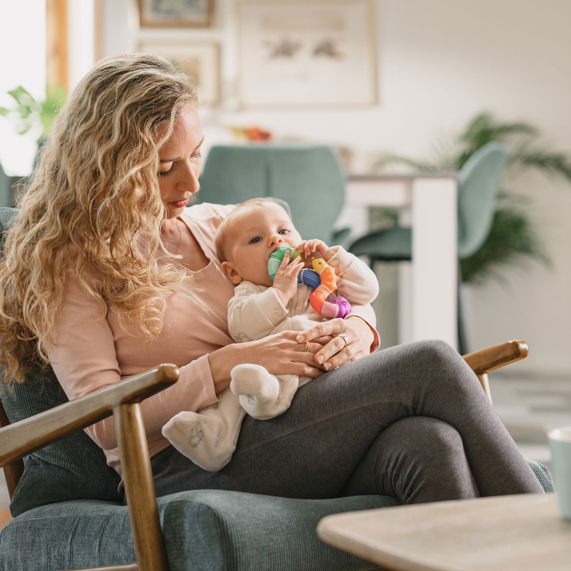 Woman holding a child in her lap. The child is teething, chewing on a Nuby Twista Rattle Teether Toy.